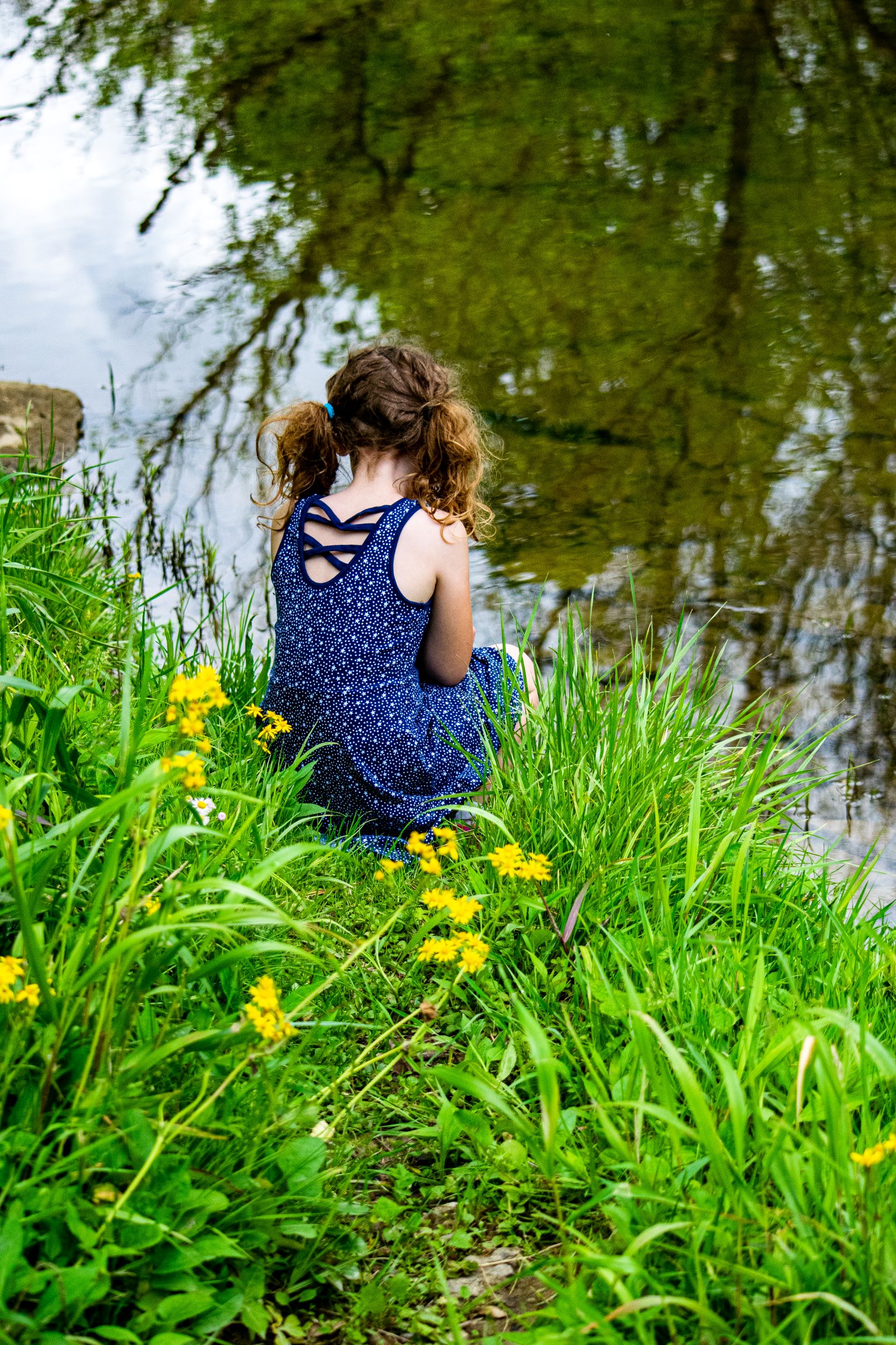 a young girl, with her back to the camera, sits at the edge of a pond on green grass